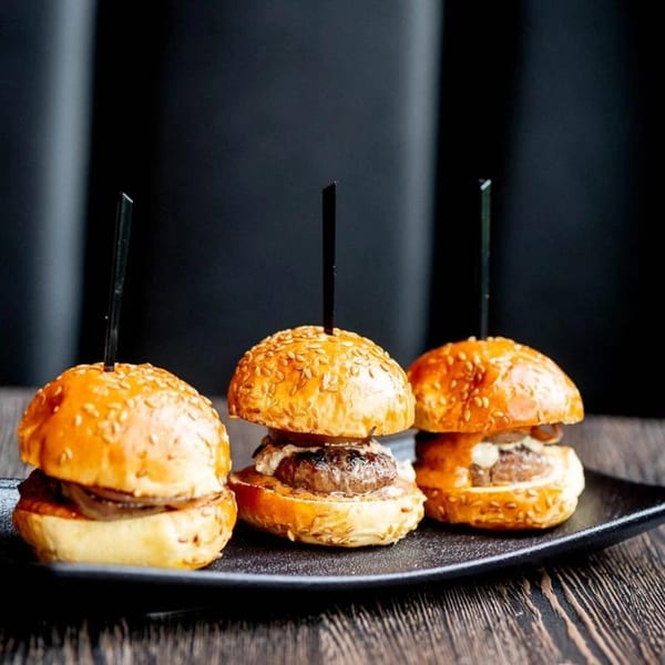 Three Craft Kitchen + Bar sliders with sesame seed buns and dark skewers on a black plate.