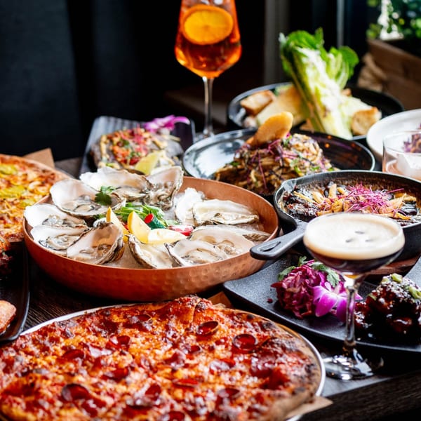 A table laden with various dishes including oysters, pizza, salad, and cocktails at Craft Kitchen + Bar.