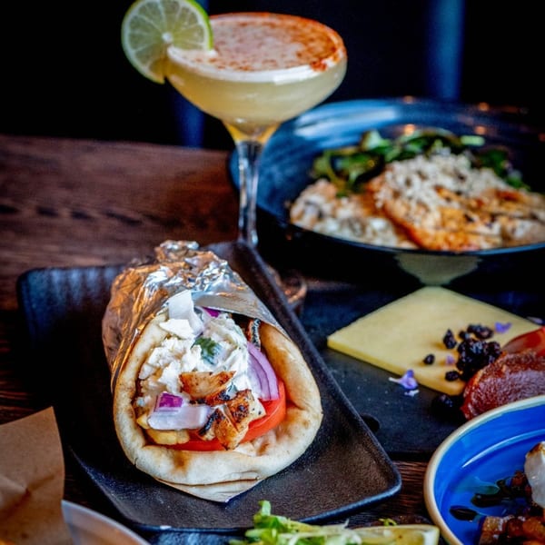 A chicken gyro, cocktail, and other dishes on a dark wooden table at Craft Kitchen + Bar.