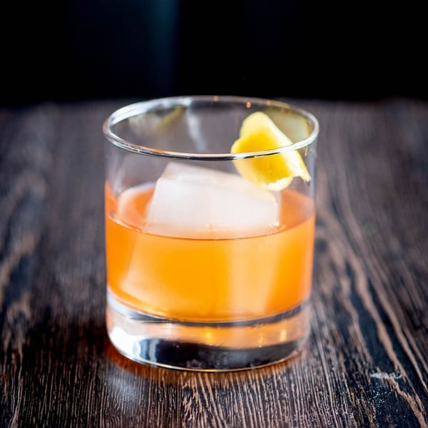 Craft Kitchen + Bar's amber cocktail with a large ice cube and lemon peel on a dark wooden table.